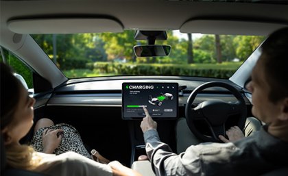 A photo taken inside an EV showing a driver and passenger in the front seat, the driver is pointing to the vehicle's charging screen.